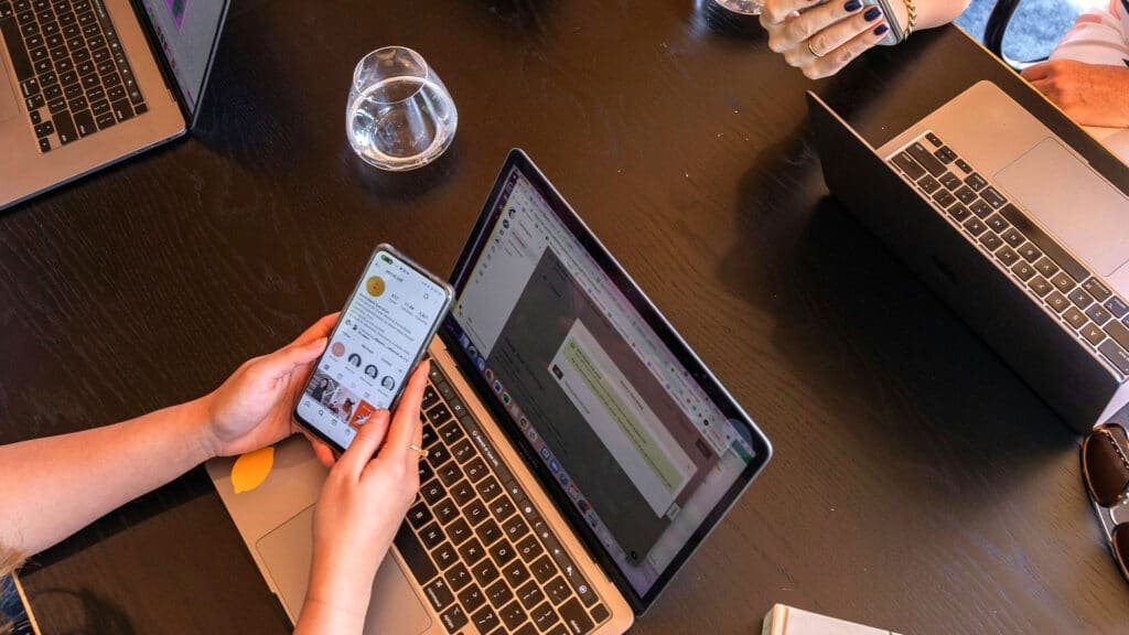 Bir masa üzerinde dizüstü bilgisayarlar arasında oturan biri, akıllı telefonunda sosyal medya profiline bakıyor.