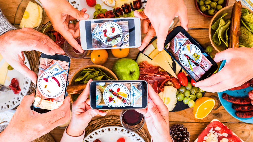 Bu görsel üstten çekilmiş bir yemek masasının her bir köşesinden akıllı telefonları ile yemeklerin fotoğrafını çeken 4 farklı el görüyoruz.