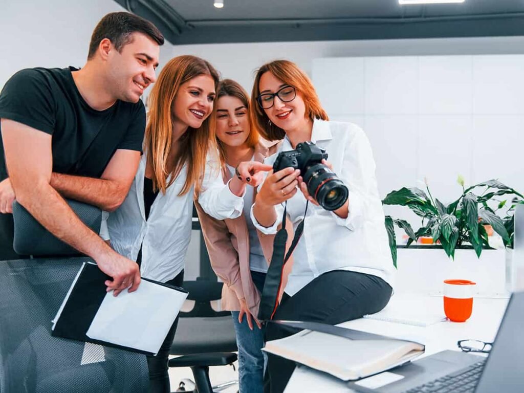 bu fotoğrafta sosyal medya yönetimi yapan reklam ajansında çalışan farklı mesleklere sahip insanlar masanın etrafında fotoğraf makinesinden fotoğrafa baktığı görülüyor