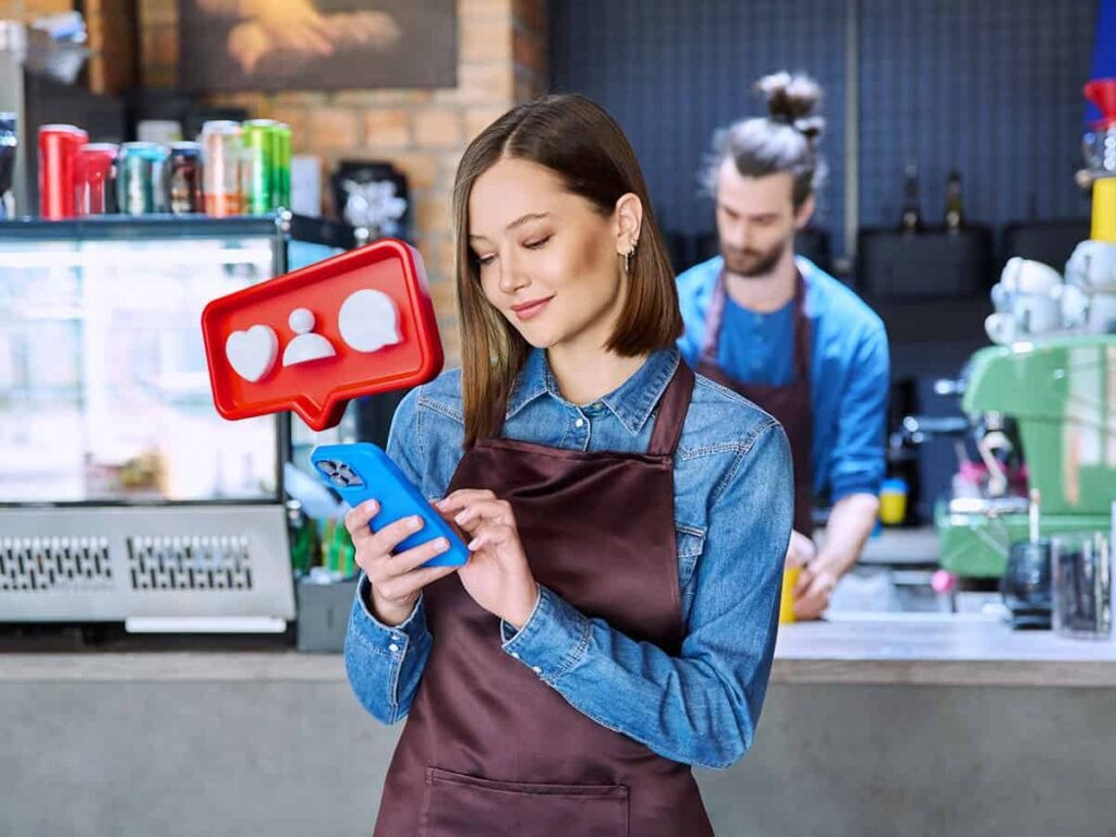 Bu fotoğrafta sosyal medya yönetimi yapan küçük işletme sahibinin gelen etkileşime telefonuna bakarak gülümsediği ve arka tarafta kahve yapan erkek çalışan olduğu görüşüyor.