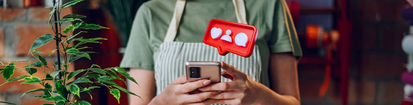 Önlük giyen bir kişi, akıllı telefonunda sosyal medya etkileşimlerini kontrol ediyor; mesaj balonunda beğeni, takipçi ve mesaj simgeleri görünüyor. Sosyal medya yönetimi, küçük işletmelerin dijital varlıklarını güçlendirme aracı olarak öne çıkıyor