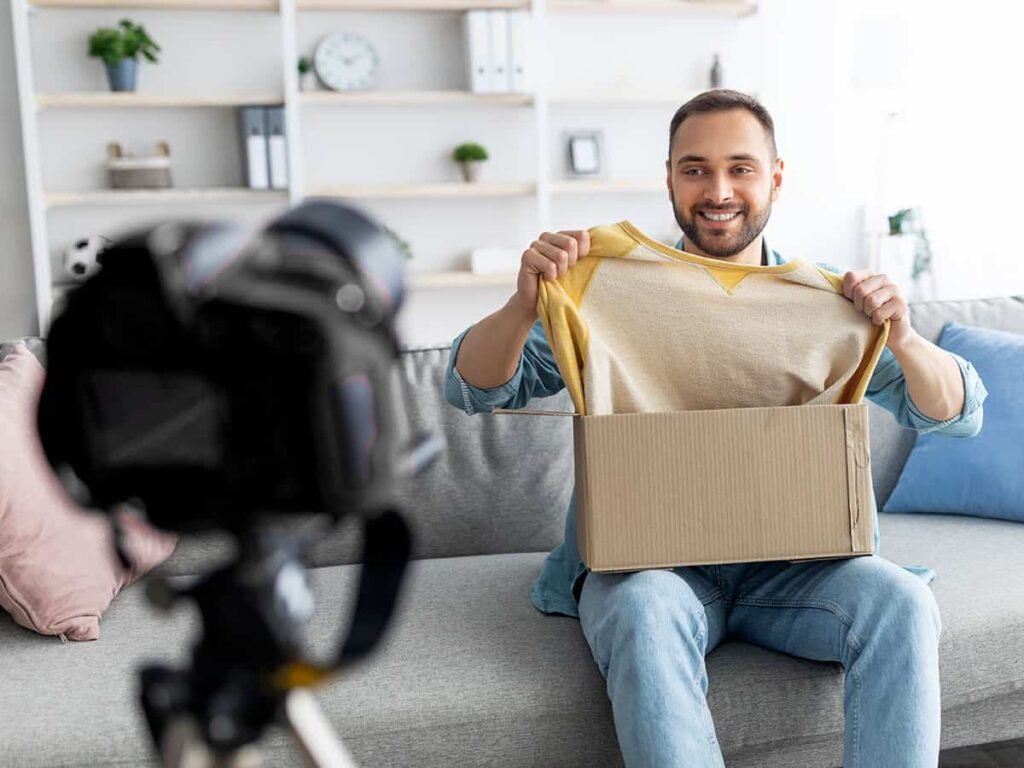 Bir erkek içerik üreticisi, ev ortamında bir kutu açılımı videosu çekiyor. Koltukta oturan adam, kutudan çıkardığı sarı kollu bir sweatshirt'ü kameraya doğru gösteriyor ve gülümsüyor. Ön planda, video kaydı yapan bir kamera bulunuyor. Arka planda ise raflar ve dekoratif eşyalar yer alıyor. Görsel, bir kutu açılımı (unboxing) videosunu ve sosyal medya içerik üretimini simgeliyor.