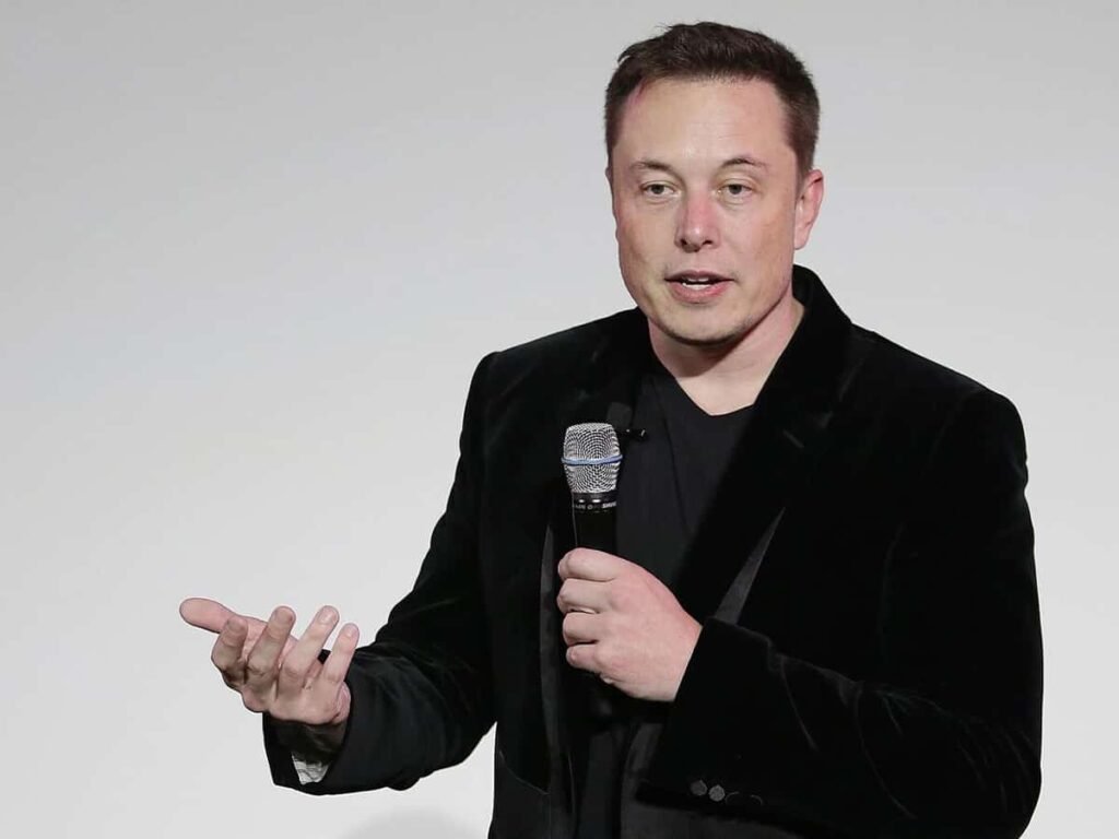 Elon Musk, mikrofon tutarak siyah bir ceketle sahnede konuşma yapıyor.