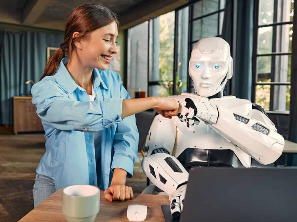 Kadın, ofis ortamında bir robotla oturmuş ve birbirlerine yumruk tokuşturuyorlar. Kadın gülümserken, robotun beyaz ve siyah parçalarla donatılmış bir yapısı var.