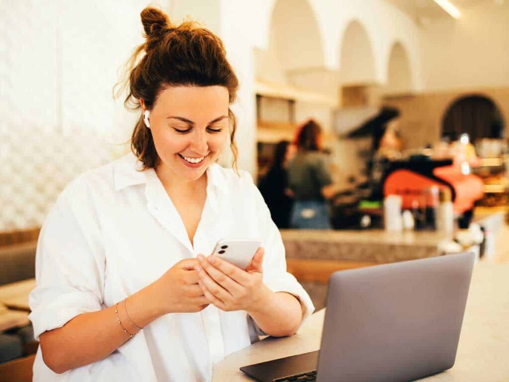 Bir kafede dizüstü bilgisayarı ile çalışan bir kadın, telefonunda mutlulukla mesaj okuyor. Arka planda hareketli bir ortam var. Bu görsel, dijital bağlantıyı, esnek çalışma tarzını ve iletişimi temsil ediyor