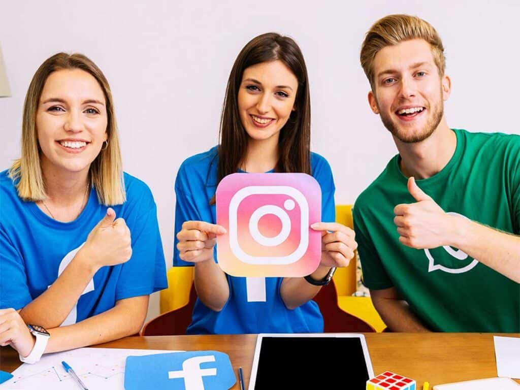 Instagram, Facebook, LinkedIn ve WhatsApp gibi sosyal medya platformlarının simgelerini masada sergileyen genç bir ekip. Ekip üyeleri, Instagram simgesini tutarken pozitif bir ifadeyle baş parmaklarıyla destek işareti yapıyor. Bu görsel, sosyal medya yönetimi ve platformlara hakimiyeti temsil ediyor, ayrıca takım ruhunu ve enerjik iş birliğini yansıtıyor