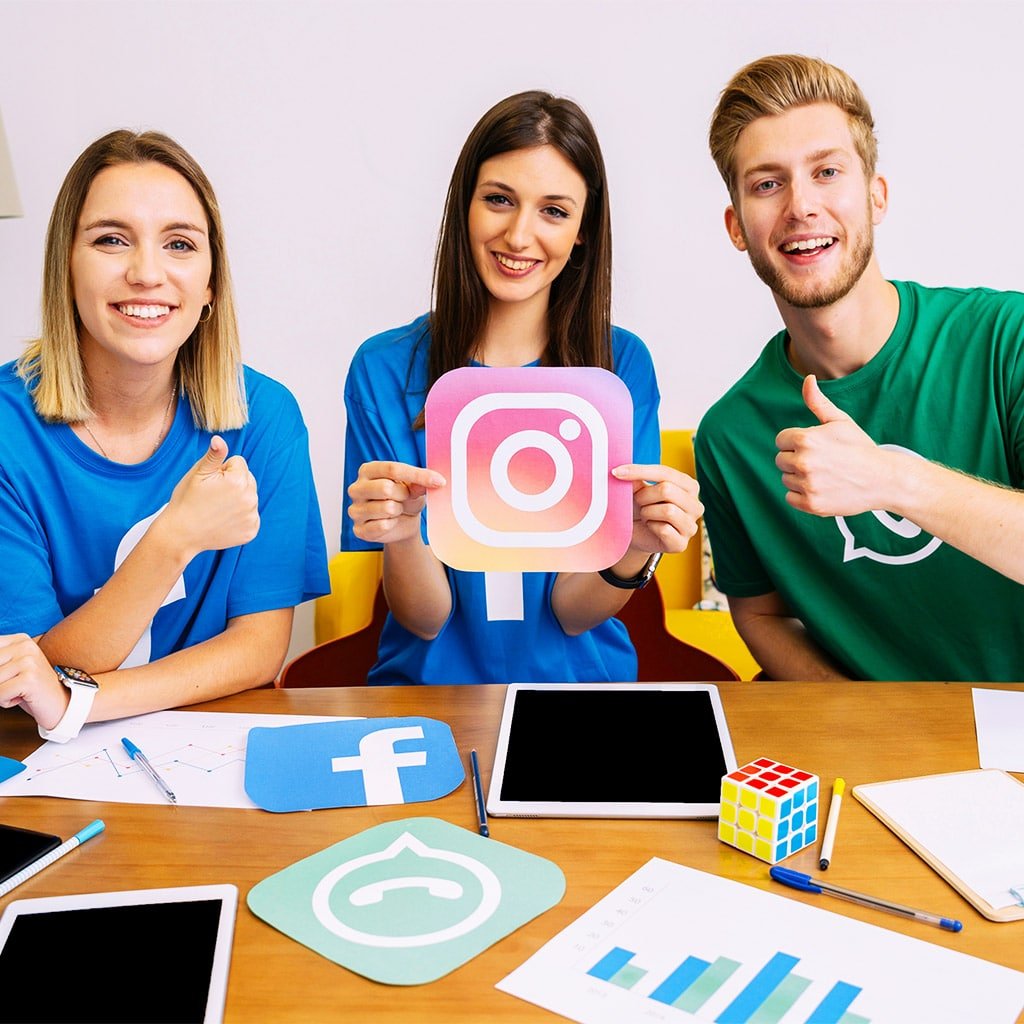 Instagram, Facebook, LinkedIn ve WhatsApp gibi sosyal medya platformlarının simgelerini masada sergileyen genç bir ekip. Ekip üyeleri, Instagram simgesini tutarken pozitif bir ifadeyle baş parmaklarıyla destek işareti yapıyor. Bu görsel, sosyal medya yönetimi ve platformlara hakimiyeti temsil ediyor, ayrıca takım ruhunu ve enerjik iş birliğini yansıtıyor