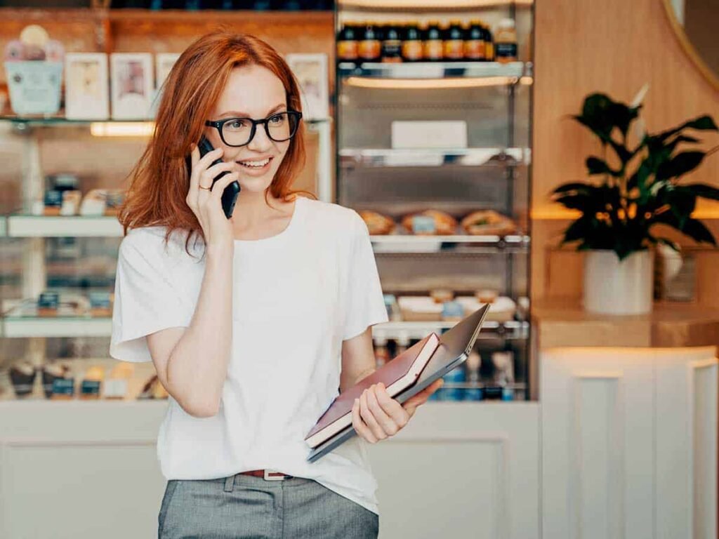Telefonla konuşan, elinde defter ve dizüstü bilgisayar taşıyan gözlüklü bir kadın. Arkasında bir kafe vitrini var, iş dünyasında aktif ve enerjik bir profesyonel imajı yaratıyor.