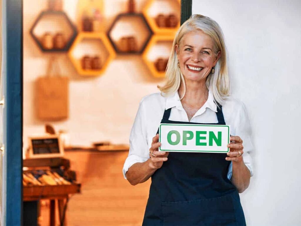 Kadın elinde 'Open' tabelasıyla.Yerel bir işletme sahibi, mağazasını açmanın heyecanını yansıtırken sıcak bir gülümsemeyle karşınızda.